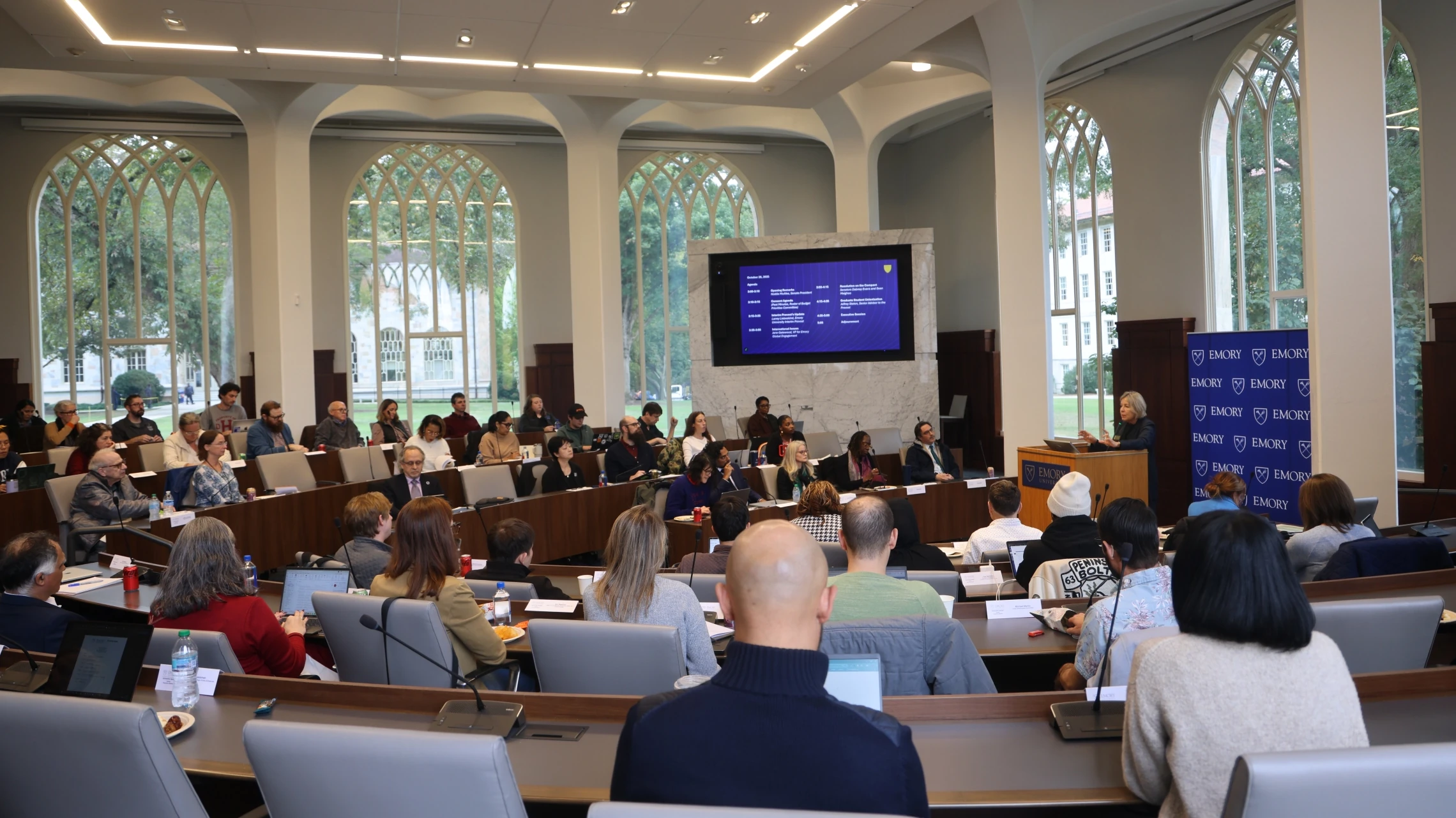 Wide view of the Emory University Senate meeting hall with gothic arched windows, attendees seated, and a presentation displayed on screen